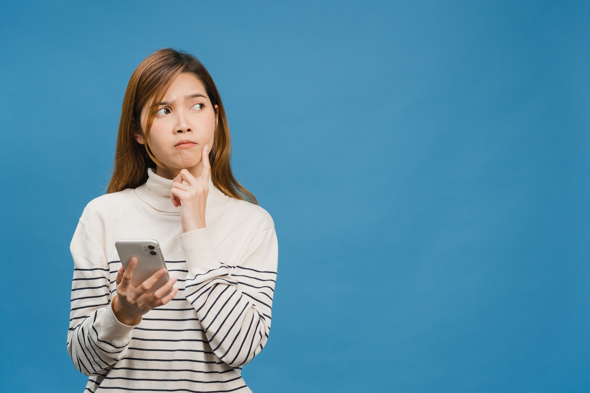Thinking dreaming young Asia lady using phone with positive expression, dressed in casual clothing feeling happiness and stand isolated on blue background. Happy adorable glad woman rejoices success.